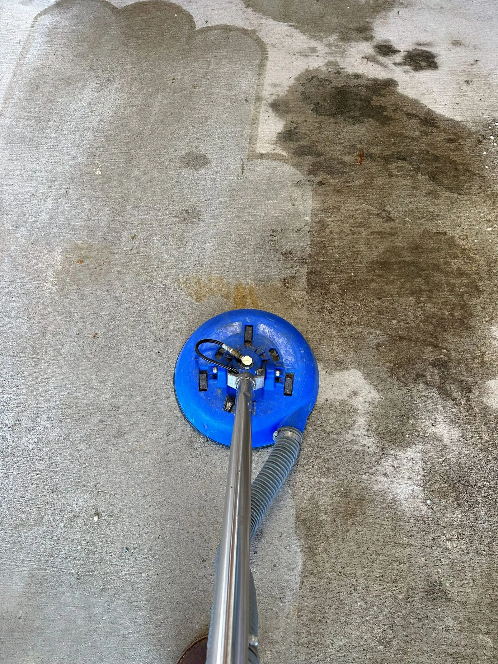 A rotary hard-surface cleaning tool part way across a concrete floor, with a sharp line between the cleaned and soiled halves